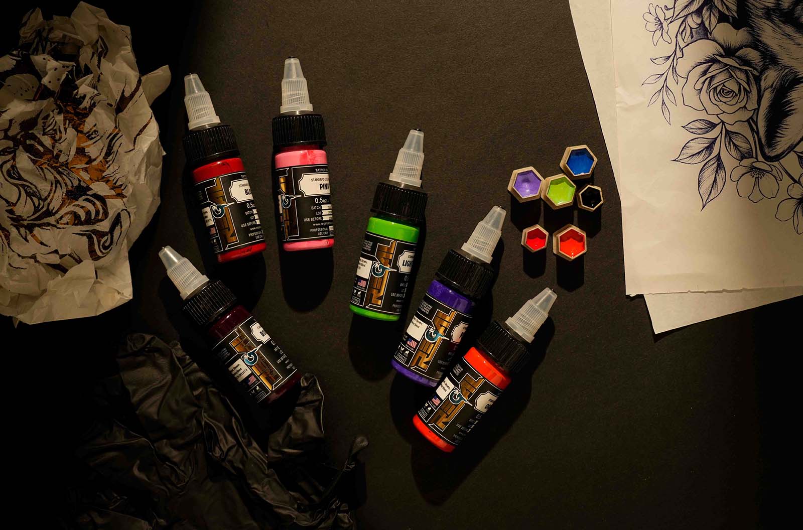 Regal tattoo ink bottles and color caps arranged beside a floral tattoo stencil design on a dark workstation.